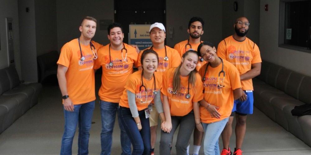 Image of a diverse group of students wearing matching orange t-shirts that say "Special Olympics MedFest," smiling and posing for a group photo indoors