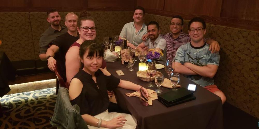 Image of a diverse group of eight people in their twenties, sitting at a booth in a restaurant with glasses of water, turning to smile at the photographer.